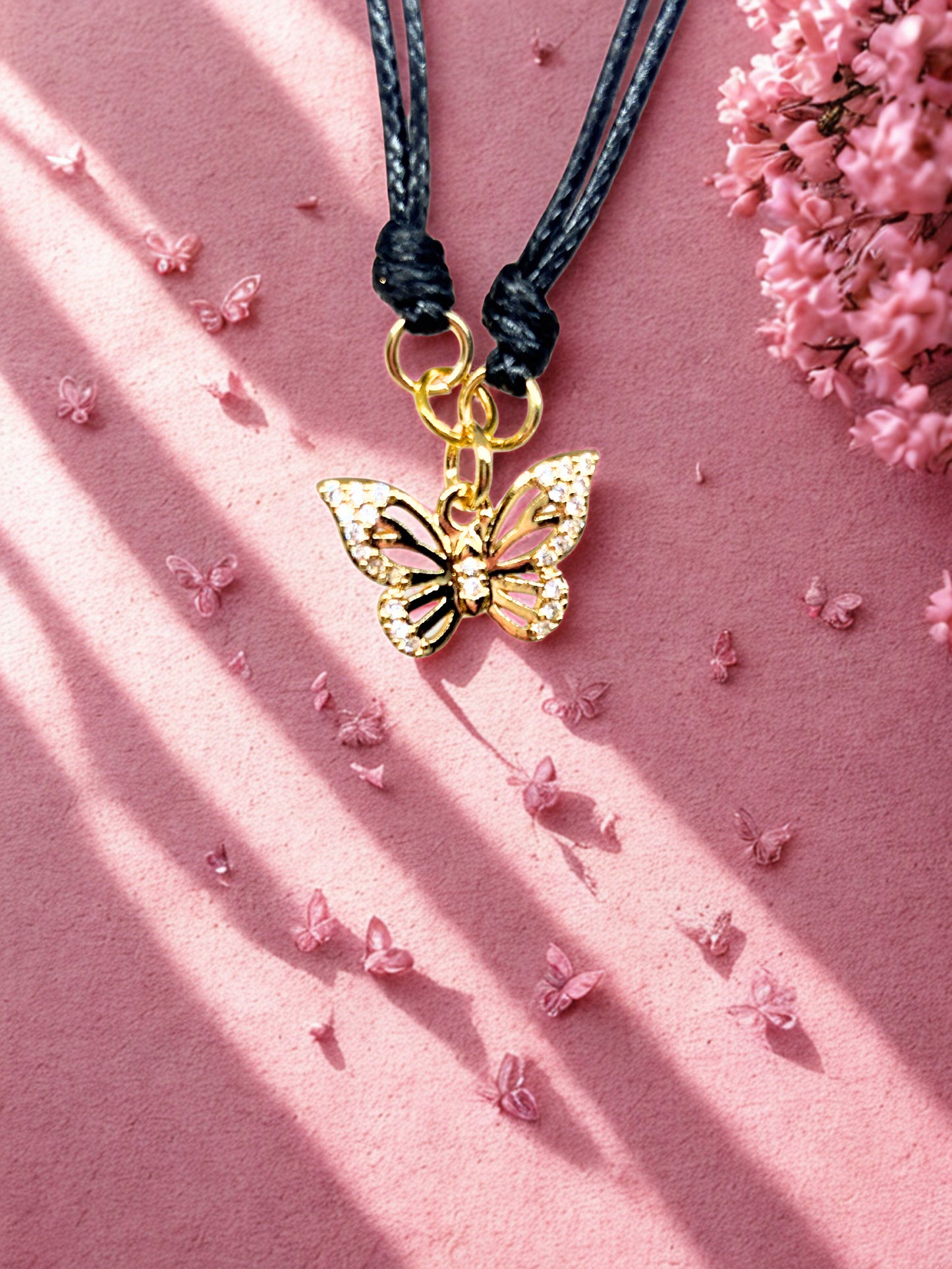 Gold butterfly charm on a black cord against a pink background with small flowers.