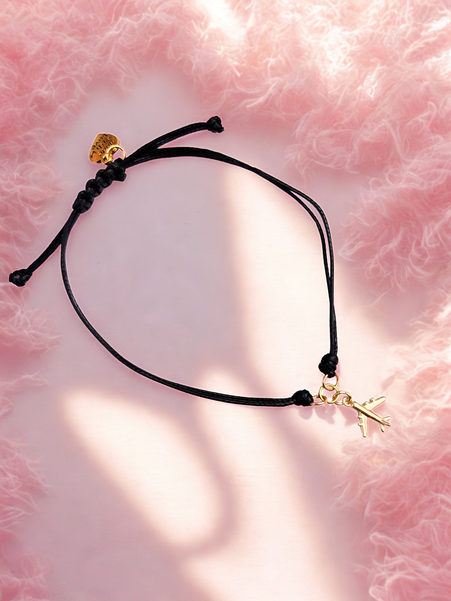 Black cord bracelet with gold aeroplane charm on a pink textured background