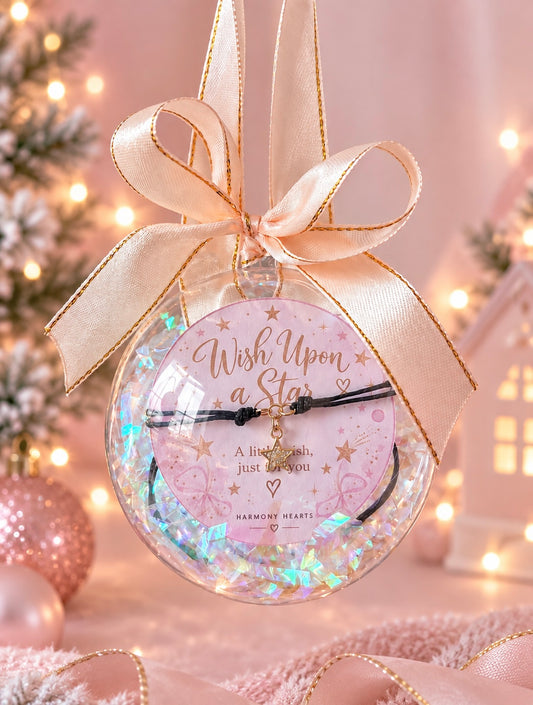 A round clear ornament with iridescent shred a pink circular insert reading 'Wish Upon a Star,' and a small gold star charm bracelet, tied with a champagne ribbon in a softly lit holiday setting.