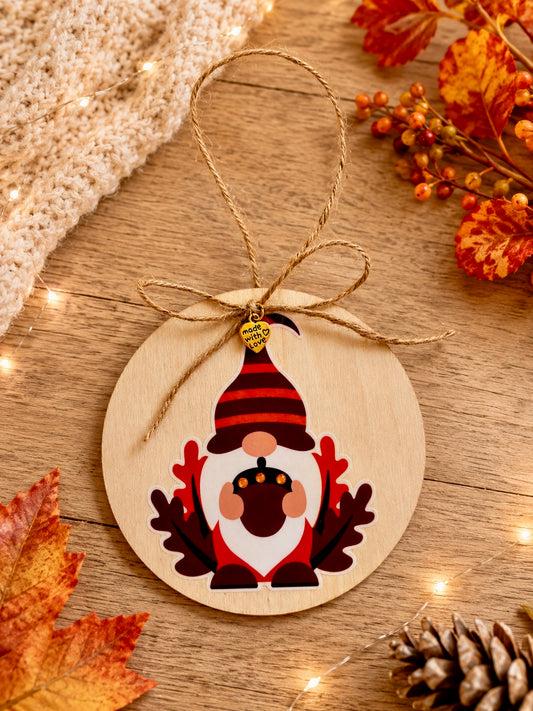 A round wooden hanging ornament featuring a festive gnome design with a red striped hat, white beard, and acorn motif, suspended by a twine loop on a rustic wood surface with autumn leaves and string lights.