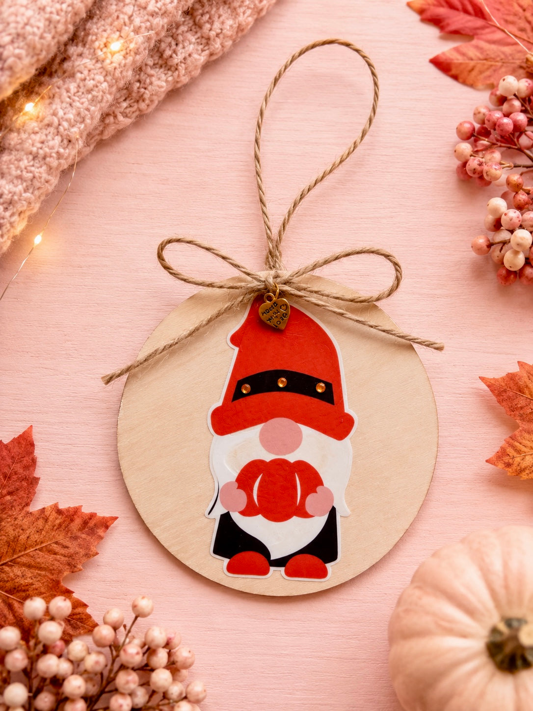 A round wooden ornament tag with a hanging twine loop and bow, featuring a printed gnome illustration in red, white, and black. The ornament is styled on a pink surface with autumn leaves, berries, a small pumpkin, and a knitted textile.