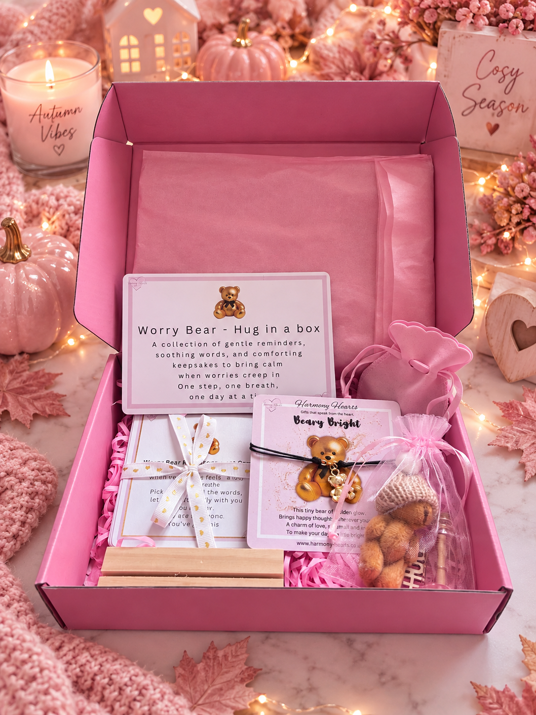 A pink gift box filled with cozy items, including a card reading 'Worry Bear - Hug in a box,' a wooden block, a teddy bear charm bracelet. The box is styled on a marble surface with pink knit fabric, candles, and autumn decorations in the background.