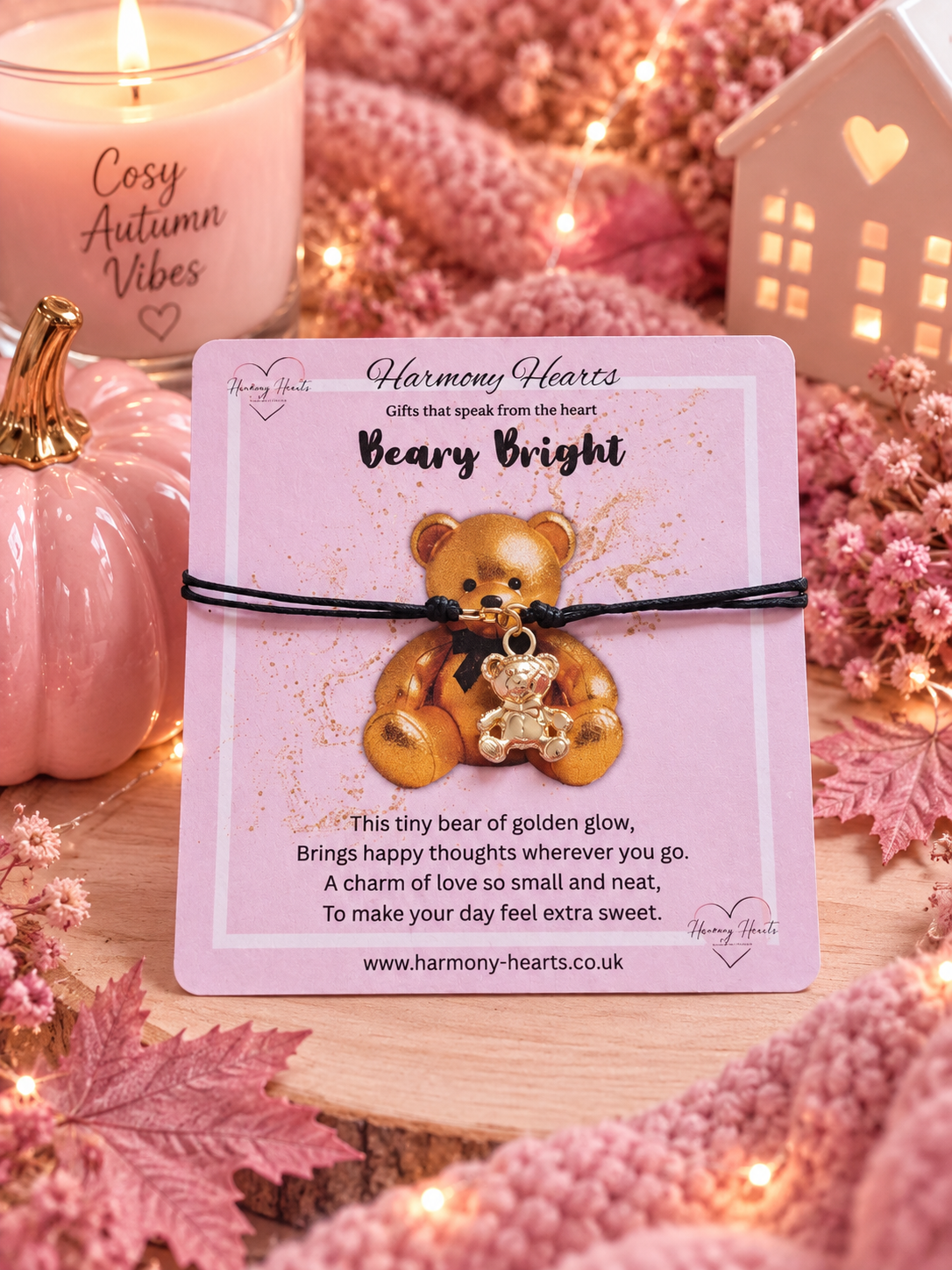 A gold teddy bear charm bracelet on a black cord, displayed on a pink card labeled 'Harmony Hearts Beary Bright'. The card is styled on a wooden surface with pink autumn leaves, a lit candle, a small pink pumpkin, and a warm white house ornament in the background.