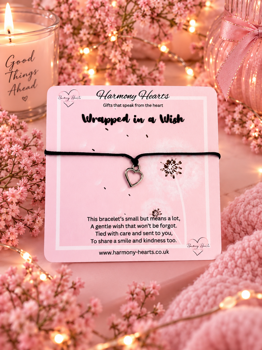 A small black cord bracelet with a heart-shaped charm and a silver floral accent, displayed on a pink card labeled Harmony Hearts. The card is styled with pink flowers, a lit candle, and warm fairy lights in the background.